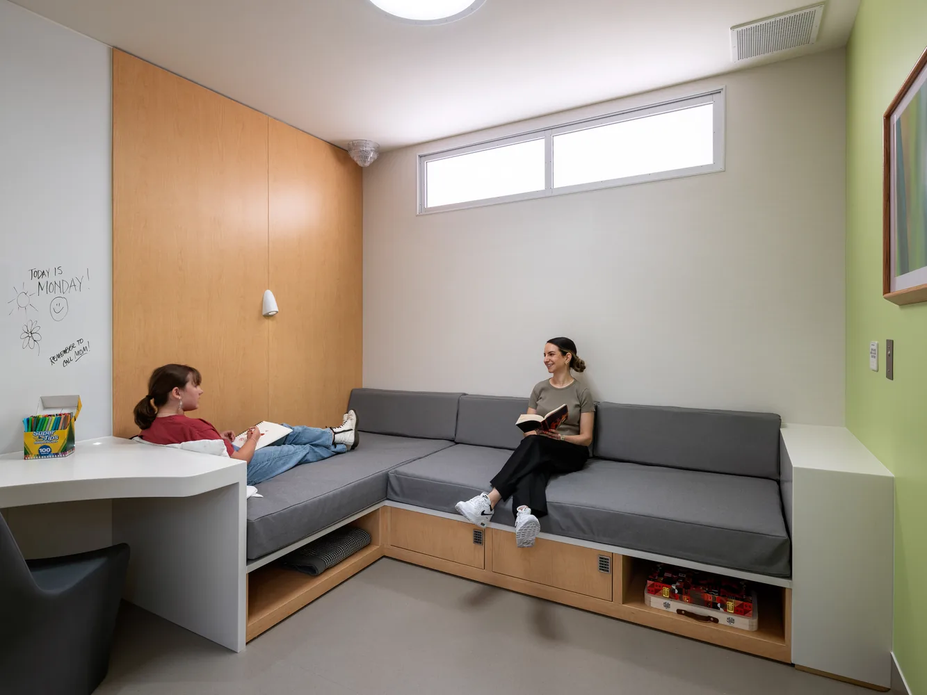 Two young people sit and read on a built-in upholstered bench in a bedroom with a maple wardrobe, a clerestory window and a writable wall carrying a hand-drawn note.